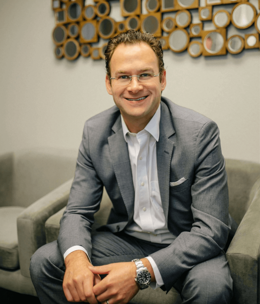 Professional man in a gray suit sitting on a gray couch, smiling confidently. The background features a modern decorative wall with circular mirrors, creating a contemporary office atmosphere. This image conveys professionalism and approachability, suitable for business-related content. Ideal for use on a corporate website or personal brand profile to enhance engagement and establish credibility.