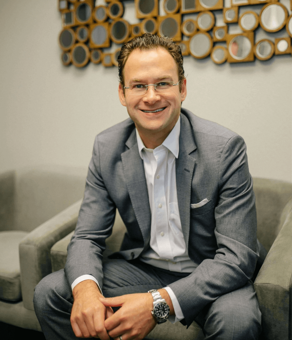 Professional man in a gray suit sitting on a gray couch, smiling confidently. The background features a modern decorative wall with circular mirrors, creating a contemporary office atmosphere. This image conveys professionalism and approachability, suitable for business-related content. Ideal for use on a corporate website or personal brand profile to enhance engagement and establish credibility.