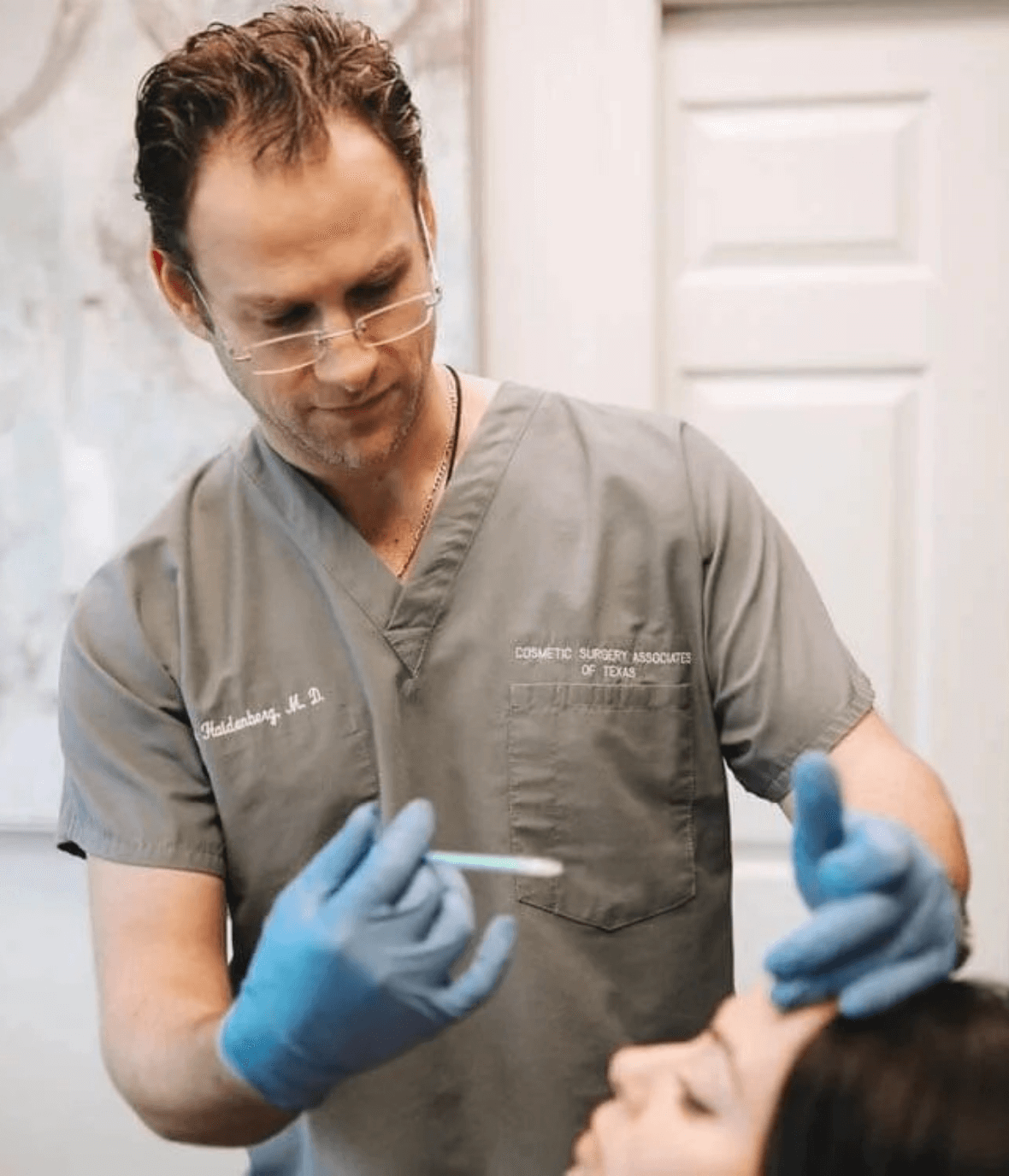 A medical professional in gray scrubs and blue gloves performs a cosmetic procedure on a patient, focusing on facial aesthetics. The clinician holds a syringe, demonstrating expertise in administering injections, likely for dermal fillers or Botox. The clinical setting features a clean, modern background, emphasizing a professional environment for cosmetic treatments. This image highlights the practice of aesthetic medicine, showcasing the careful and precise work involved in enhancing facial features safely and effectively.