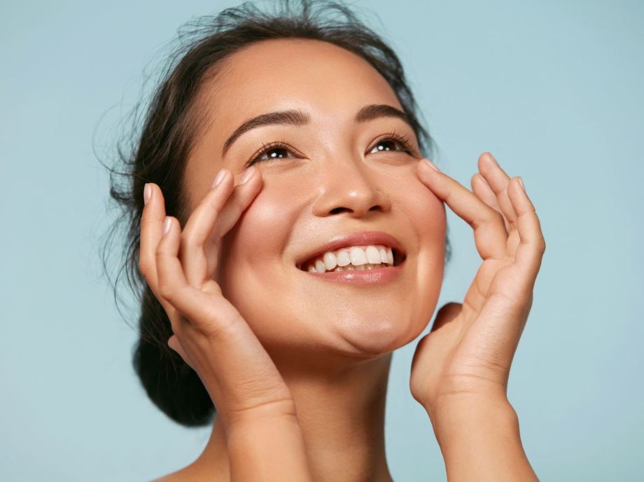 A young woman with a radiant smile gently applies skincare around her eyes, showcasing a healthy and glowing complexion. The background features a soft blue hue, enhancing the fresh and vibrant look. This image embodies self-care and beauty, highlighting the importance of skincare routines for maintaining youthful skin. The woman’s expression conveys happiness and confidence, making it an inspiring representation of personal wellness and beauty practices.
