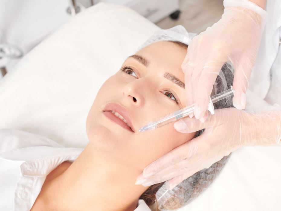 A patient receiving a cosmetic treatment, such as dermal filler injections, while lying on an examination table. A medical professional, wearing gloves, is carefully administering the treatment near the patient's lips. The setting appears clean and clinical, emphasizing a professional environment for aesthetic procedures. This image highlights the process of enhancing facial features through non-surgical techniques, commonly sought for rejuvenation and volume restoration.