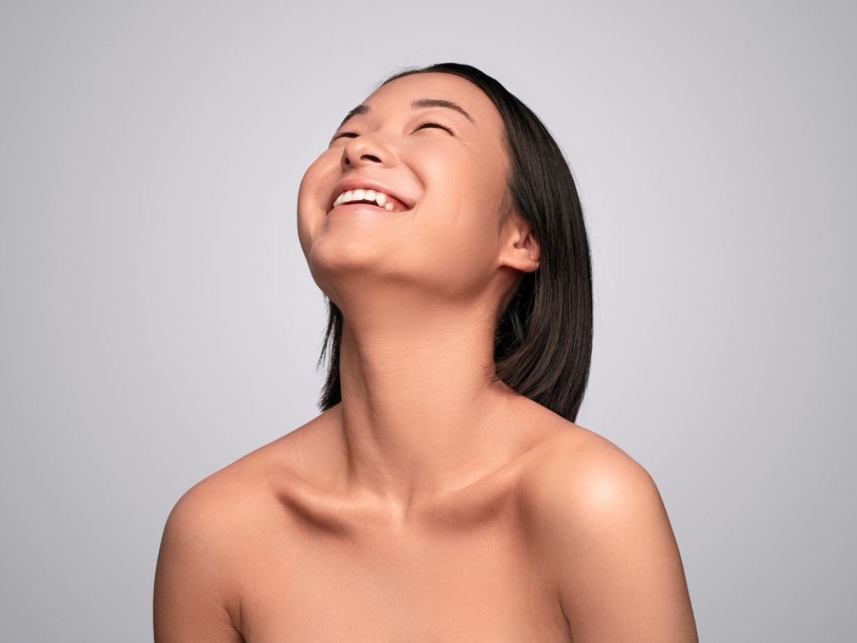 A young woman with short black hair is smiling joyfully while looking upward against a soft gray background. Her skin appears radiant and healthy, emphasizing a natural beauty. The image captures a moment of happiness and positivity, showcasing the subject's expressive features and warm demeanor. This portrait reflects themes of joy and self-confidence, making it ideal for discussions around beauty, wellness, and personal growth.