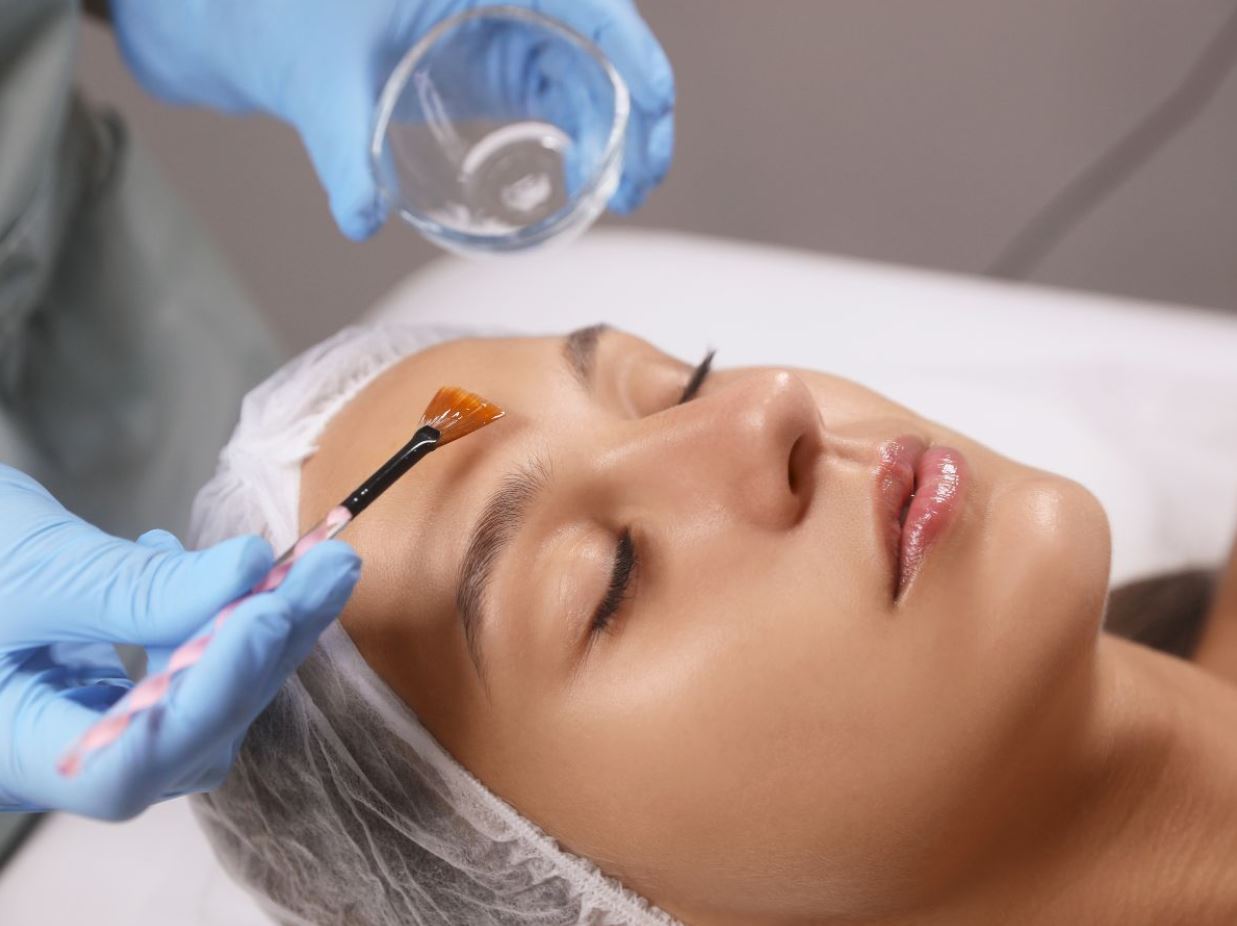 Aesthetic treatment in progress, featuring a woman receiving a facial application. A beautician, wearing blue gloves, uses a brush to apply a product to the woman's forehead. The woman has her eyes closed and appears relaxed, with a hairnet covering her hair. A small glass bowl with product is visible in the background. This image highlights the skincare process, emphasizing professional beauty care and relaxation.