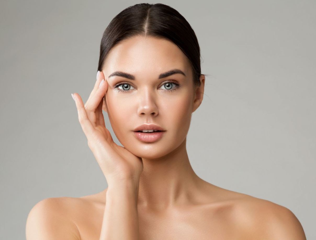 Close-up of a woman with smooth, radiant skin, showcasing a natural beauty look. She has long, dark hair pulled back, highlighting her facial features. The woman is gently touching her face with one hand while maintaining direct eye contact with the camera. The soft gray background enhances her fresh, glowing complexion. This image can be used in beauty and skincare contexts, emphasizing skincare routines, makeup tips, or beauty product promotions. Ideal for articles or social media posts about skincare, beauty trends, or self-care