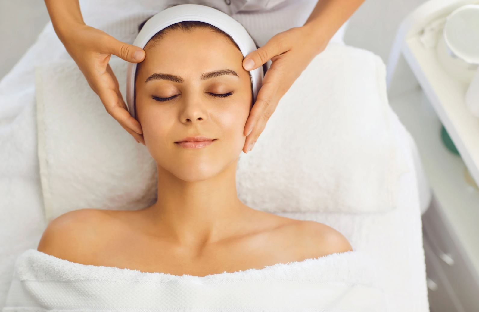 A woman receiving a relaxing facial massage at a spa. She lies on a soft towel with her eyes closed, enjoying the treatment. The therapist's hands gently cradle her face, emphasizing relaxation and self-care. The serene environment is enhanced by soft lighting and clean, minimalistic decor, creating a tranquil atmosphere. This image illustrates the benefits of spa treatments for wellness and rejuvenation.