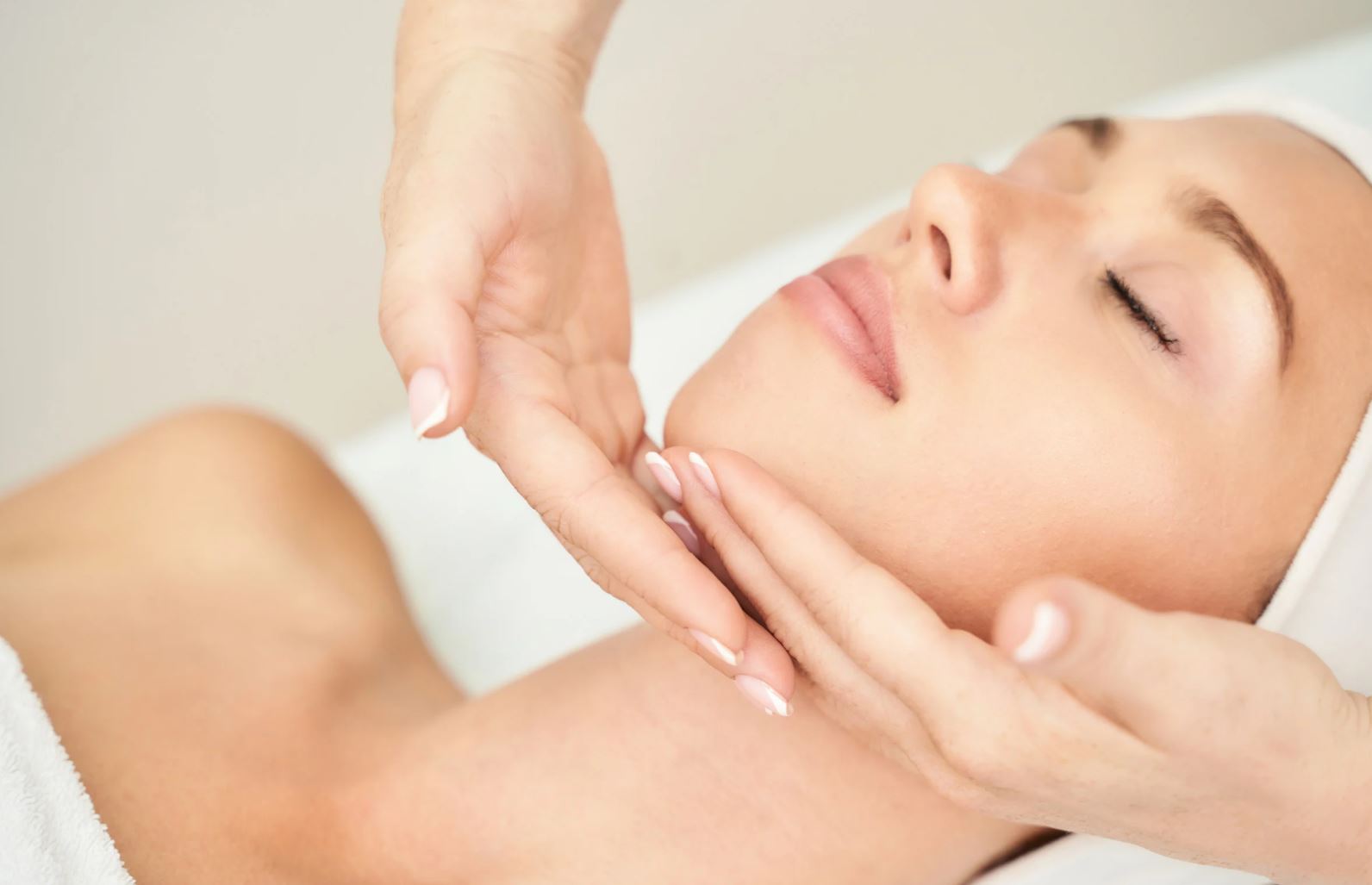 A serene woman receiving a facial treatment, with her eyes closed and a relaxed expression. A therapist's hands gently support her jawline, enhancing the soothing atmosphere of the spa environment. The woman is wrapped in a white towel, and her skin appears smooth and healthy, reflecting a professional skincare session aimed at rejuvenation and relaxation. This image represents the benefits of spa treatments and skincare routines for overall well-being.