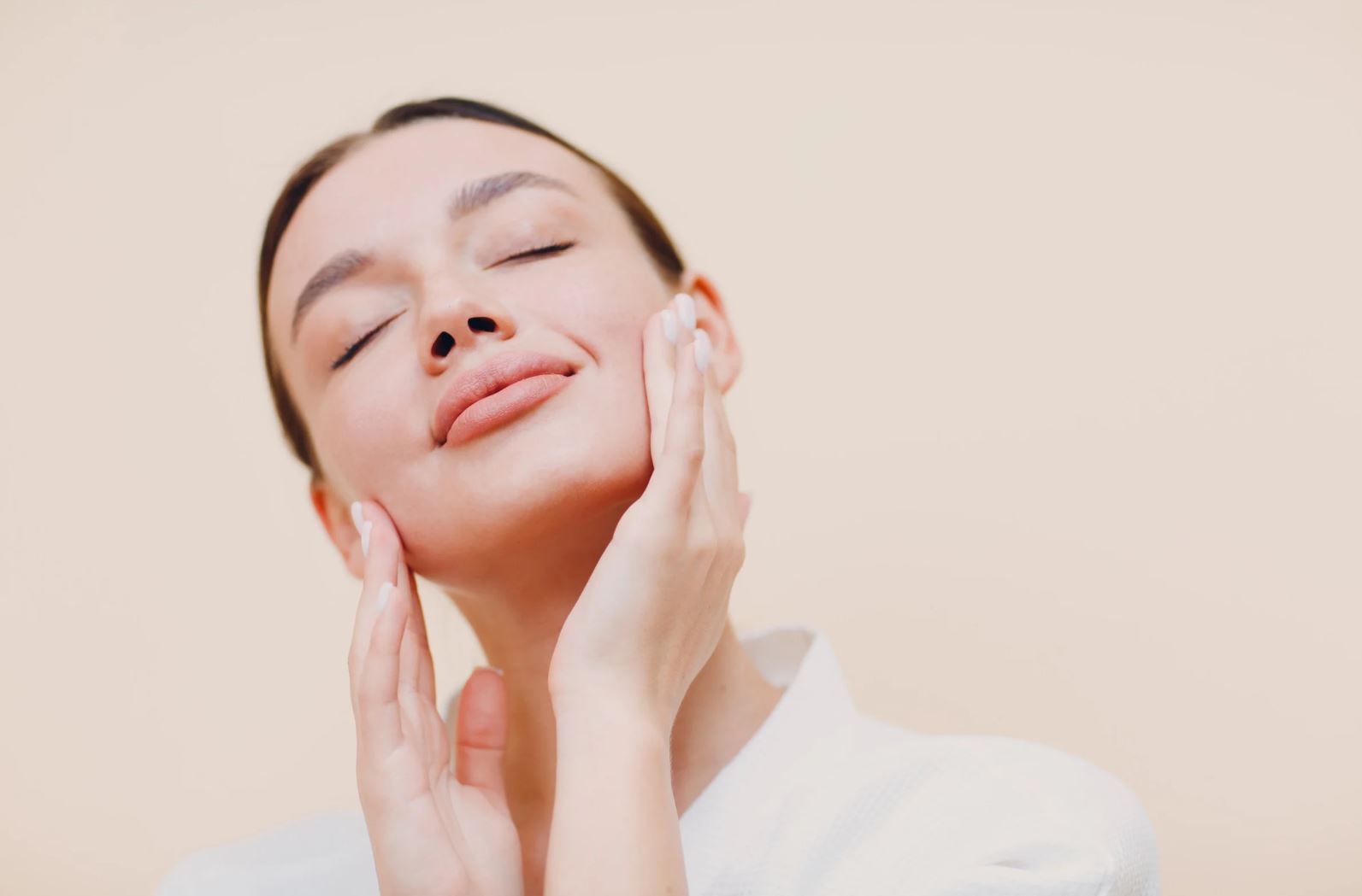 A woman with a serene expression gently applies skincare product to her face, showcasing a healthy skincare routine. She has her eyes closed, conveying relaxation and self-care. The image features a soft, neutral background that enhances the focus on her glowing skin and the act of moisturizing. This visual promotes the importance of skincare and wellness, appealing to beauty enthusiasts and those seeking effective skincare practices.