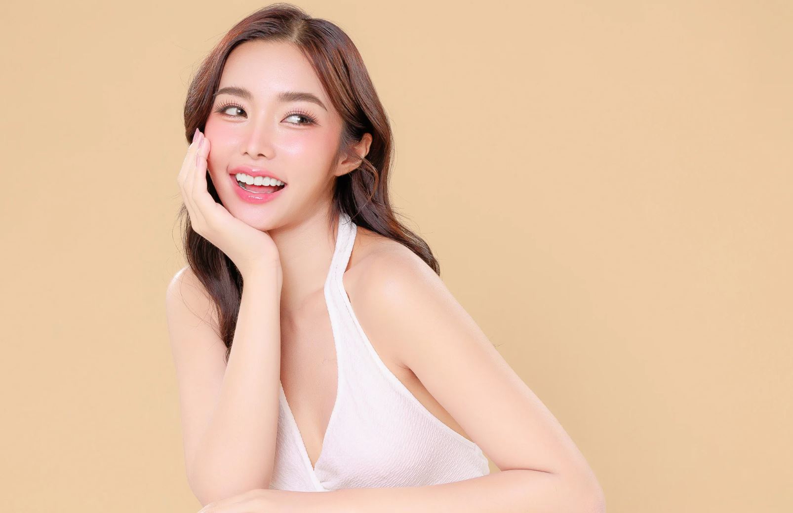 A smiling woman with long, wavy hair rests her chin on her hand against a soft, neutral background. She wears a white sleeveless top and has a natural, radiant appearance with subtle makeup. The image conveys a sense of joy and confidence, making it ideal for beauty and lifestyle content.