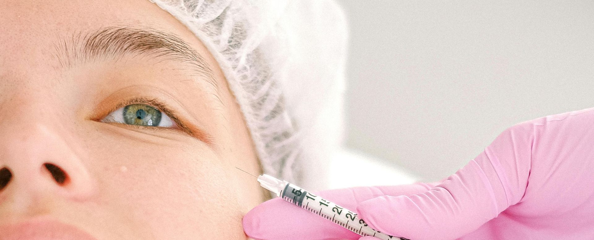 A close-up of a person's face undergoing a cosmetic injection procedure. The individual has light skin and an expression of calmness, with one eye visible and a hint of makeup. A healthcare professional, wearing pink gloves, is administering the injection with a syringe near the cheek. The background is softly blurred, emphasizing the procedure. This image represents aesthetics treatments like Botox or dermal fillers, highlighting the growing trend in non-surgical cosmetic enhancements.