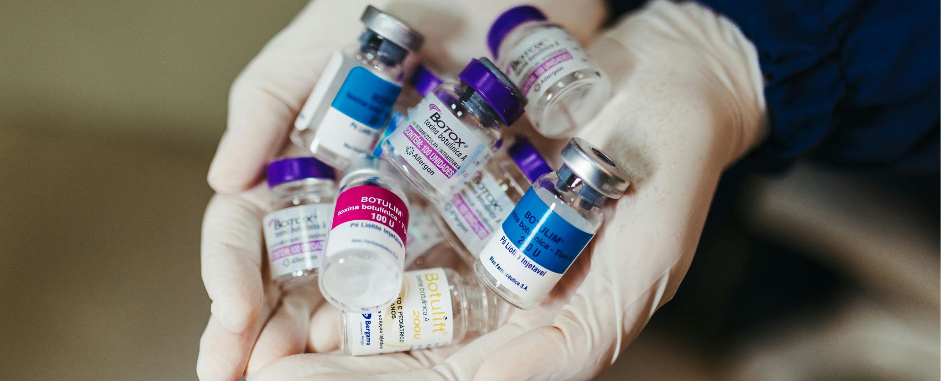 A healthcare professional wearing a glove holds a variety of botulinum toxin vials, commonly known as Botox, in their hand. The image showcases several labeled bottles, including brands like Allergan and others, highlighting their use in cosmetic and medical treatments. The background is softly blurred, emphasizing the focus on the vials. This image illustrates the preparation for Botox injections, which are popular for reducing wrinkles and treating various medical conditions, such as muscle spasms and excessive sweating.
