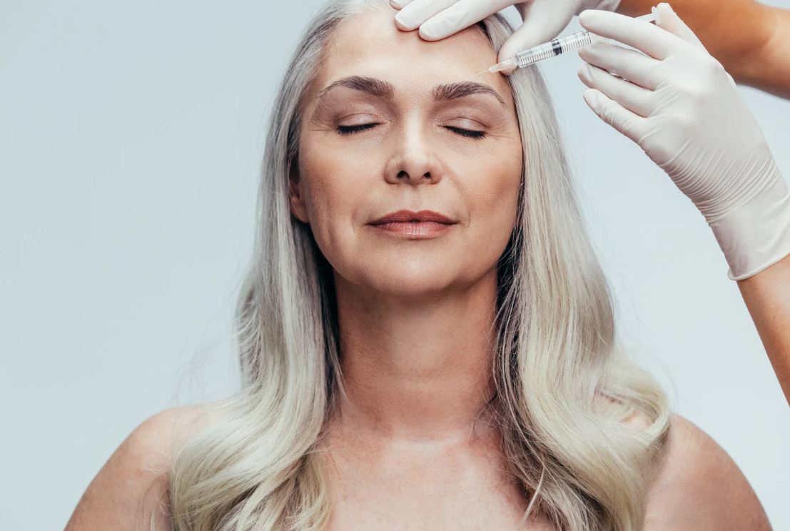 A middle-aged woman with long, straight silver hair is receiving a cosmetic treatment. A healthcare professional, wearing white gloves, is administering an injection to her forehead. The woman has her eyes closed, appearing calm and relaxed in a clinical setting. This image illustrates the process of aesthetic procedures, highlighting the importance of professional care in skincare and anti-aging treatments. Ideal for content related to beauty, wellness, or cosmetic dermatology.