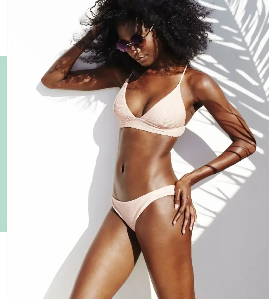 Model wearing a light pink bikini poses against a white wall, showcasing a stylish summer look. The bikini features a triangular top and matching bottoms, emphasizing a sleek silhouette. The model has voluminous, natural curls and accessorizes with trendy sunglasses, adding a chic touch. Soft shadows cast by palm leaves create a tropical ambiance, enhancing the beach-ready vibe of the image. Ideal for summer fashion inspiration, this photograph highlights body positivity and confidence in swimwear.