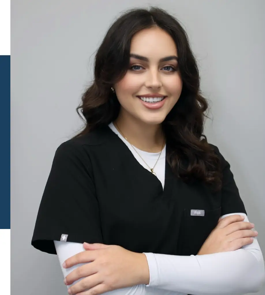 A smiling female healthcare professional wearing a black scrubs top with a white long-sleeve undershirt, standing with arms crossed. She has long, wavy hair and a subtle necklace. The background is a neutral gray, enhancing her professional appearance. This image conveys a sense of confidence and approachability, suitable for a medical or wellness-related context, emphasizing the importance of compassionate care in the healthcare industry.