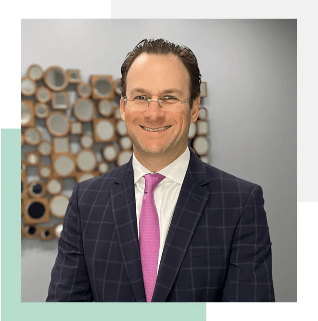 Professional headshot of a man in a dark plaid suit with a white dress shirt and a bright pink tie, smiling confidently. The background features an artistic arrangement of circular mirrors in various sizes. The image conveys a welcoming and professional atmosphere, suitable for business or corporate contexts. Ideal for use in profiles or promotional materials to highlight expertise and approachability.