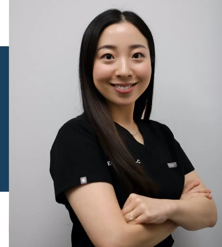 Smiling female healthcare professional wearing a black scrub top, posing with arms crossed against a light gray background. The image conveys a friendly and approachable demeanor, ideal for showcasing expertise in a medical or wellness setting. The scrub top features name tags, enhancing the professional appearance. This image is suitable for use on a healthcare website, promoting services or staff profiles.