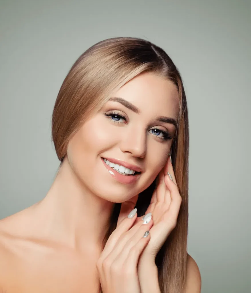 Portrait of a smiling woman with long, straight blonde hair and light blue eyes. She is gently touching her face with one hand while the other rests against her neck. The background is a soft gradient, enhancing her radiant complexion. The image conveys beauty and confidence, ideal for beauty and skincare promotions.