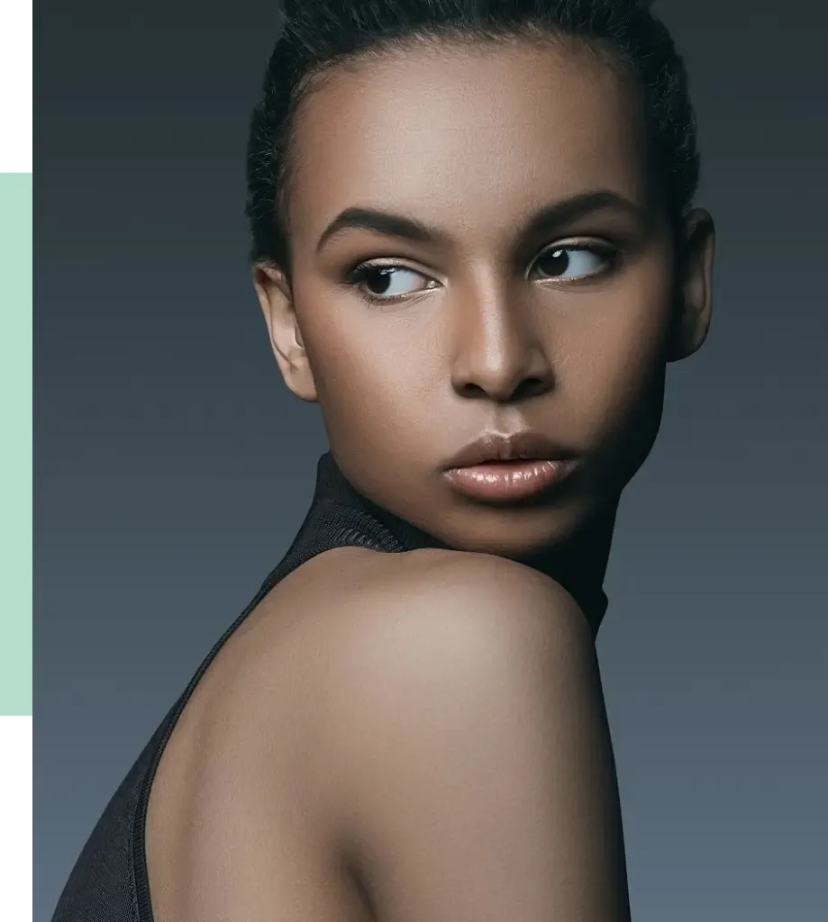 Portrait of a young woman with smooth skin and a confident expression, looking over her shoulder. She wears a sleeveless, high-neck top, highlighting her shoulder and neck. The background features a gradient from dark to light gray, enhancing her features. The image captures modern beauty and elegance, making it ideal for fashion or beauty-related content.