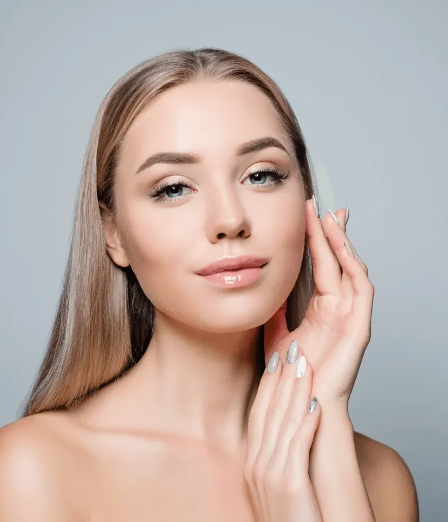 A young woman with long, straight hair poses confidently, gently touching her face with one hand. She has a natural makeup look, featuring subtle eye makeup and glossy lips, which enhances her radiant complexion. Her nails are elegantly manicured, showcasing a silver design. The background is softly blurred in light tones, creating a serene atmosphere that highlights her beauty. This image embodies themes of skincare, beauty routines, and self-care, appealing to audiences interested in cosmetics and wellness.