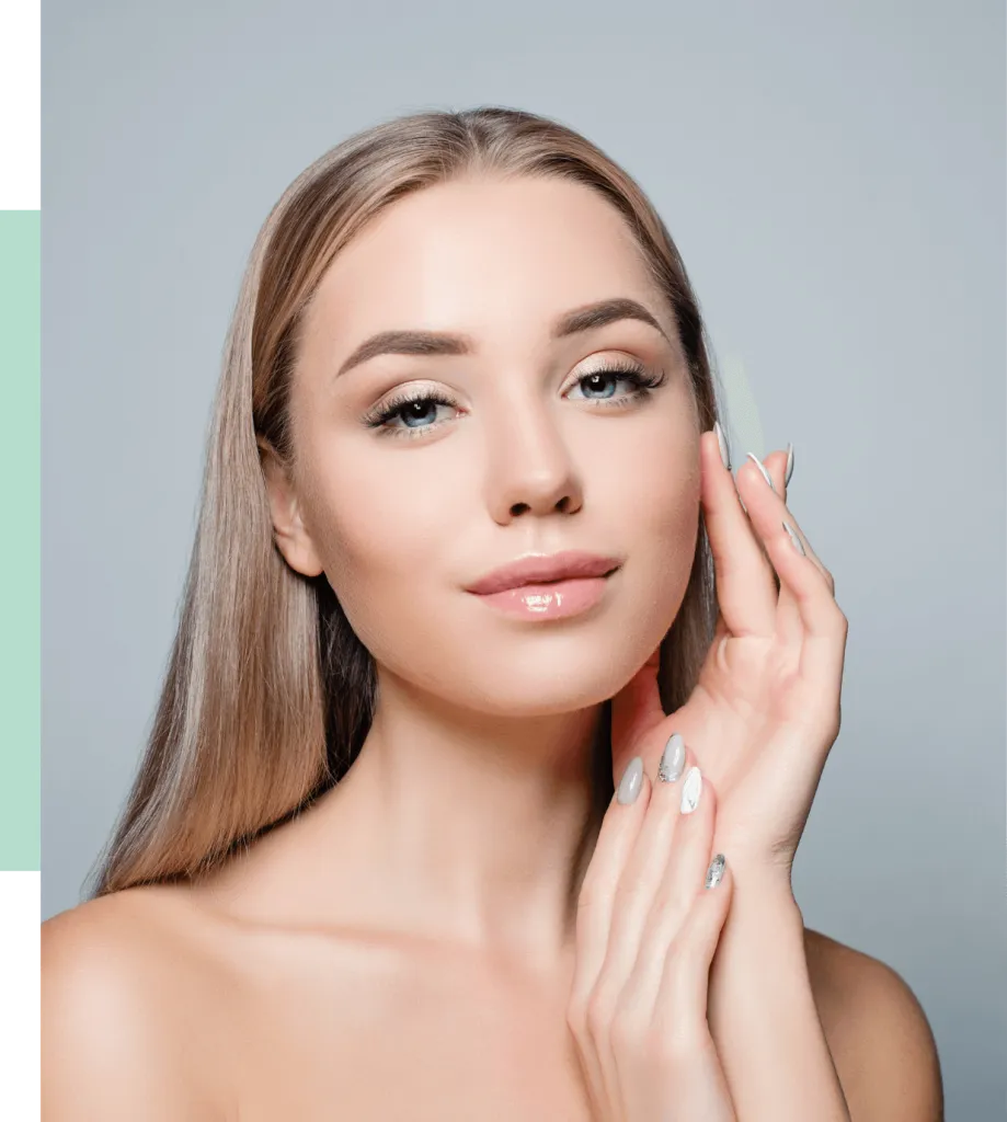 A young woman with long, straight hair poses confidently, gently touching her face with one hand. She has a natural makeup look, featuring subtle eye makeup and glossy lips, which enhances her radiant complexion. Her nails are elegantly manicured, showcasing a silver design. The background is softly blurred in light tones, creating a serene atmosphere that highlights her beauty. This image embodies themes of skincare, beauty routines, and self-care, appealing to audiences interested in cosmetics and wellness.