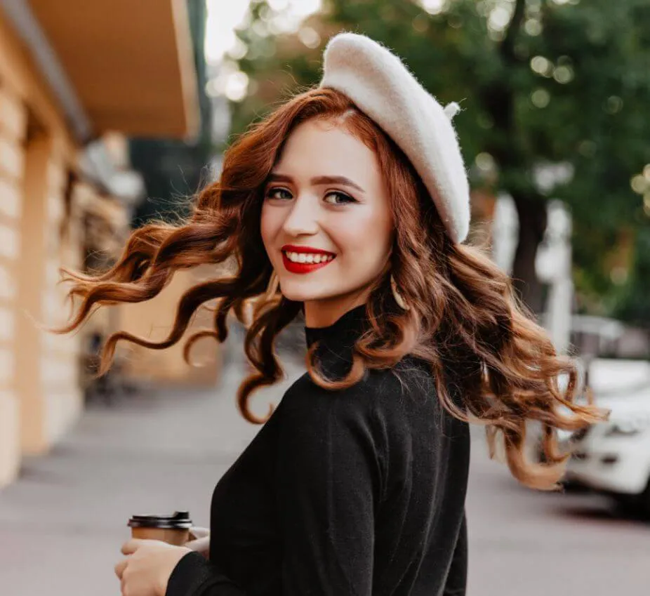 A young woman with wavy, shoulder-length hair and a white beret smiles joyfully while holding a coffee cup. She wears a black sweater and stands on a city street with blurred buildings and greenery in the background. Her bright red lipstick enhances her cheerful expression, making her the focal point of the image. The scene captures a lively urban atmosphere, ideal for showcasing fashion and lifestyle content.