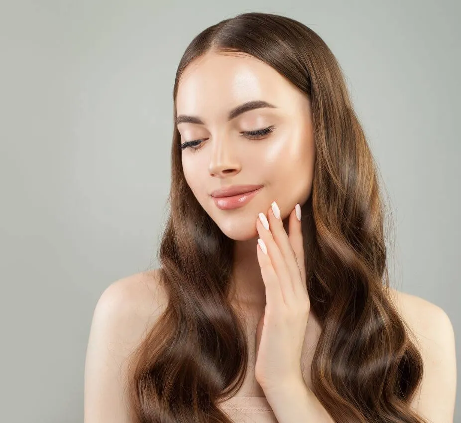 A young woman with long, wavy brown hair gently touches her face, showcasing a natural and radiant makeup look. She has a soft smile and her skin appears smooth and glowing against a light gray background. The image highlights her elegant style and emphasizes beauty and skincare. This portrait is ideal for beauty blogs, skincare product promotions, or hairstyle inspiration.