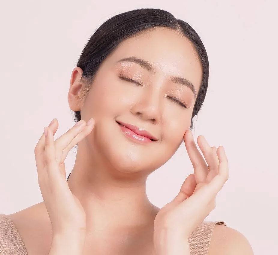 Close-up portrait of a woman with smooth skin and a radiant smile, gently touching her face with both hands. She has her eyes closed, suggesting relaxation and enjoyment. The background is soft and neutral, enhancing the focus on her glowing complexion. This image conveys a sense of beauty and skincare, ideal for promoting skincare products or beauty services.