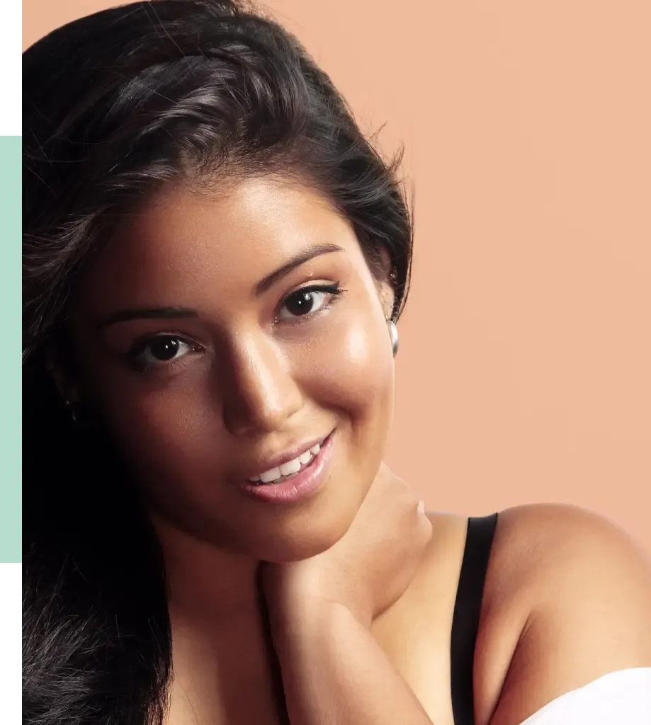 Portrait of a smiling woman with long dark hair, wearing a black top. She has a warm expression, with one hand resting on her neck. The background features a soft peach color, enhancing her natural beauty. This image captures a moment of confidence and approachability, ideal for beauty or lifestyle content.
