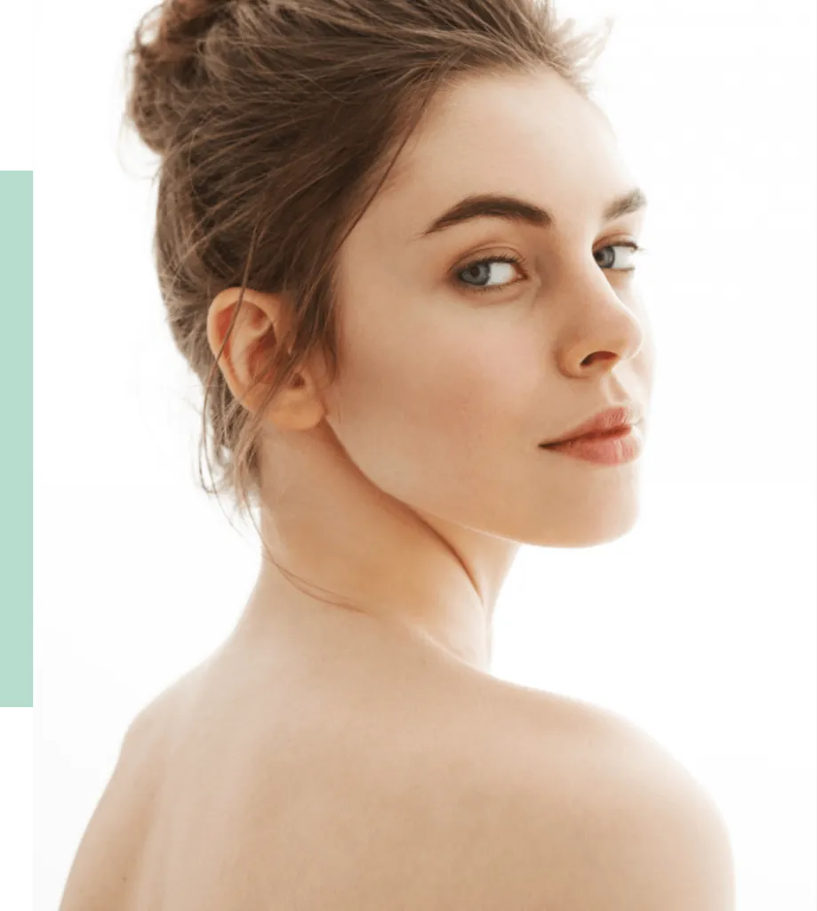 A young woman with long, brown hair styled in a bun gazes over her shoulder, showcasing her natural beauty. The image features a soft, neutral background that highlights her clear skin and expressive eyes. This portrait emphasizes a minimalist aesthetic, making it suitable for beauty and skincare content. The serene expression and gentle lighting create a calming atmosphere, ideal for promoting wellness and self-care themes.