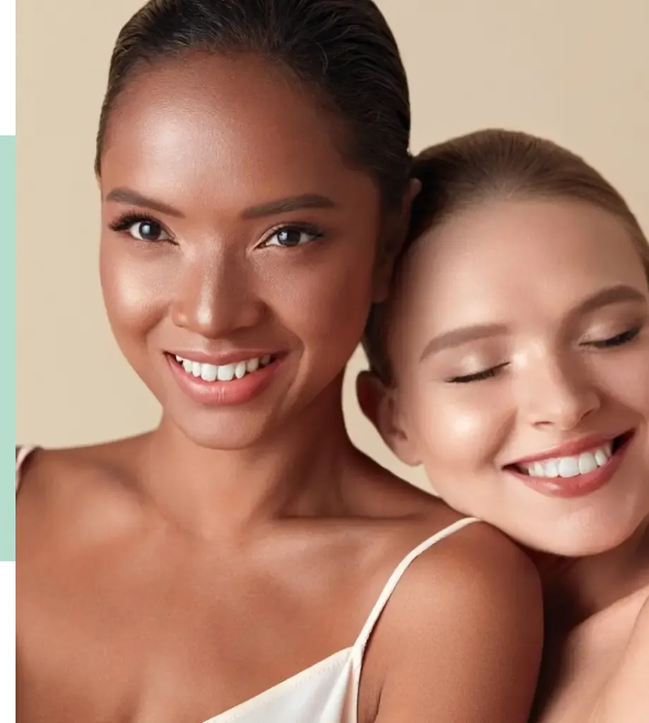 Two women with glowing skin and bright smiles pose closely together against a soft beige background. The woman on the left has dark hair and a radiant complexion, while the woman on the right has light hair and a joyful expression. Both are wearing minimal makeup, highlighting their natural beauty. This image captures a moment of friendship and confidence, showcasing diverse beauty and positivity. Ideal for content related to skincare, beauty products, or empowerment.
