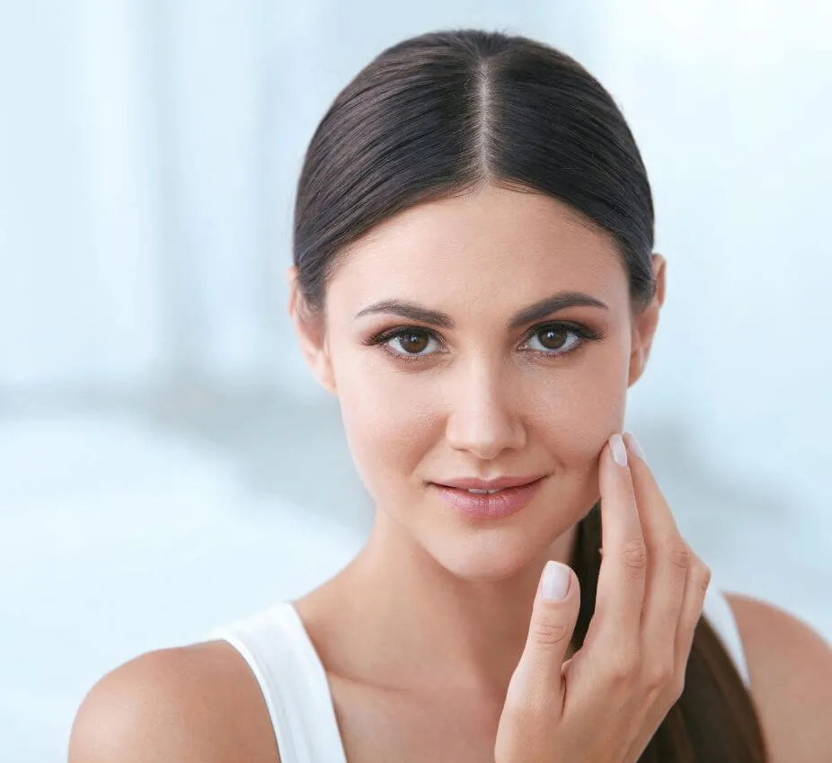 A young woman with long, dark hair and natural makeup gently touches her cheek while smiling. She is wearing a white sleeveless top and is set against a soft, blurred background. This image highlights healthy skin and a confident expression, making it ideal for topics related to skincare, beauty, or wellness.