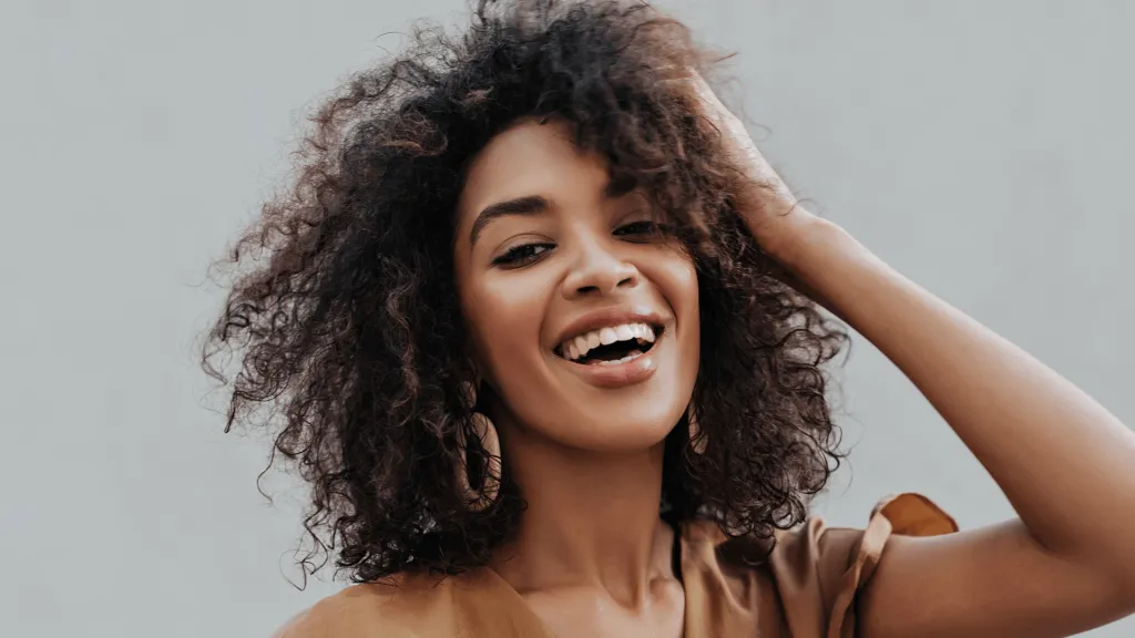 Smiling woman with curly hair poses playfully, showcasing her natural beauty. She wears a stylish brown top and large earrings, with a light gray background that highlights her joyful expression. The image captures a sense of confidence and warmth, making it ideal for content related to fashion, beauty, or lifestyle.