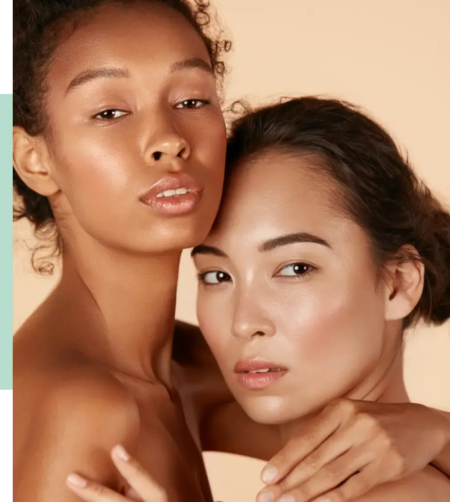 Two women with diverse skin tones pose closely together against a soft, neutral background. They have natural makeup highlighting their features, showcasing a radiant and healthy complexion. Their expressions convey confidence and connection, emphasizing beauty diversity. The image promotes inclusivity and self-acceptance, making it ideal for beauty or skincare brands.