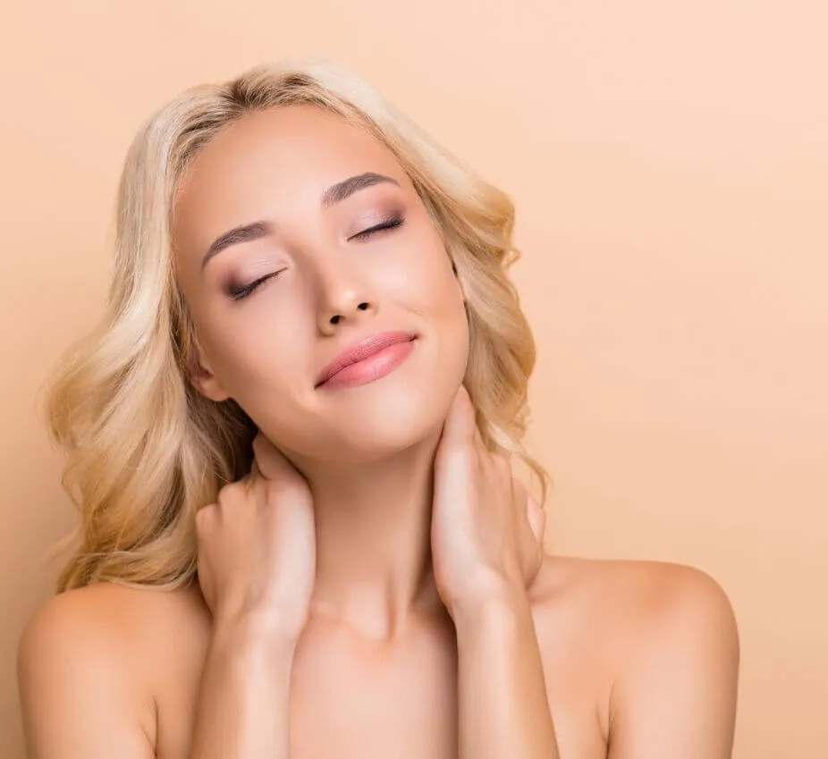 A serene young woman with long, wavy blonde hair gently touches her neck while smiling softly. She has closed her eyes, exuding a sense of relaxation and tranquility. The background features a warm peach tone, enhancing the peaceful ambiance of the image. This portrait captures a moment of self-care and beauty, highlighting the woman's natural radiance and confidence. Ideal for wellness and beauty-related content, this image conveys themes of relaxation, self-love, and skincare.