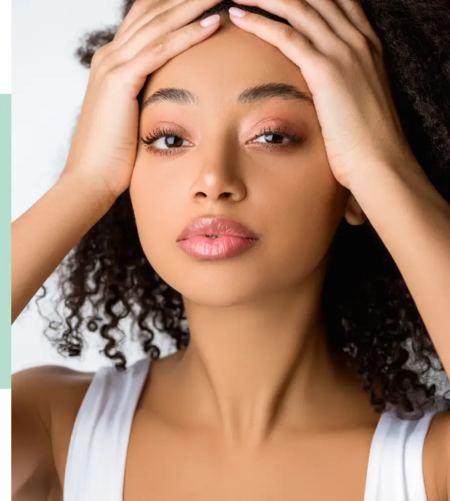 A young woman with curly hair is holding her head with both hands, displaying an expression of concern or stress. She has a natural makeup look, with soft pink tones on her lips and eyelids. The background is softly blurred, emphasizing her facial features. The image captures a moment of emotional intensity, reflecting themes of wellness and self-care.