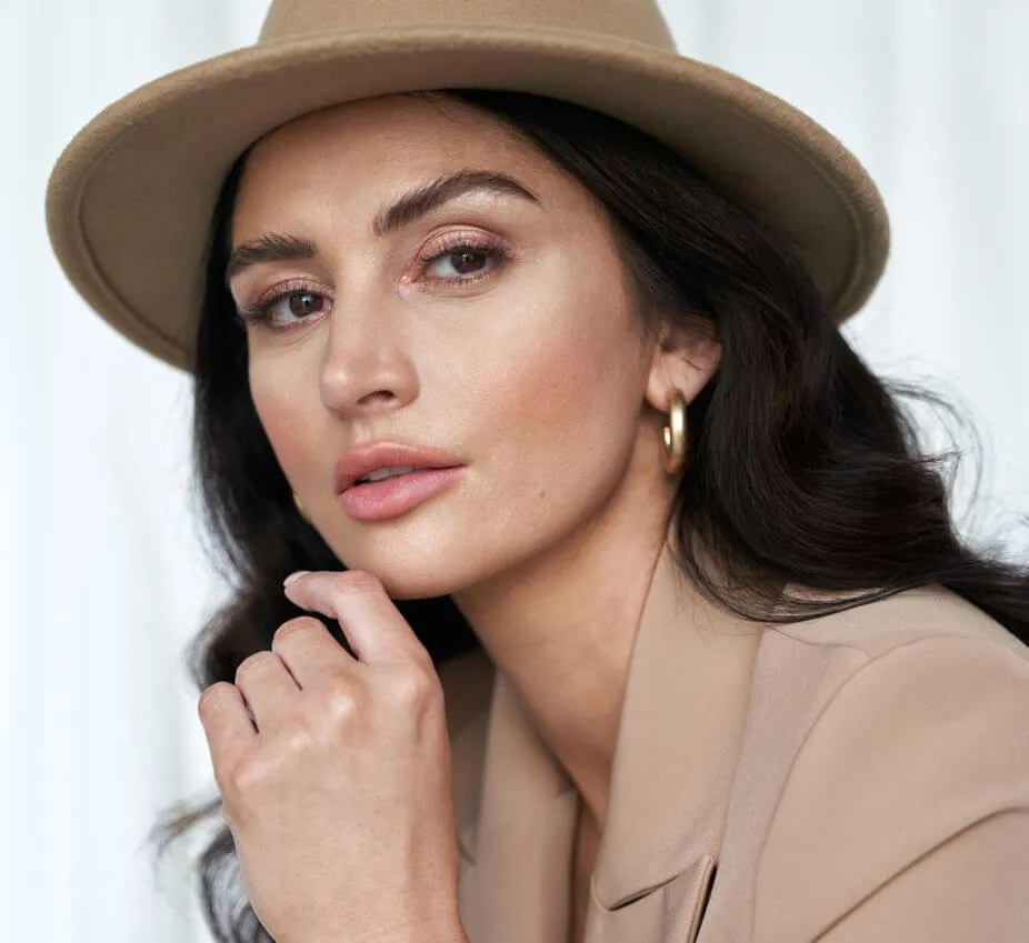 Close-up portrait of a woman with long, dark hair wearing a beige hat and gold hoop earrings. She is softly smiling, resting her chin on her hand, against a light-colored backdrop. The image highlights her natural makeup, featuring soft tones that enhance her features. This stylish look exemplifies modern fashion trends and showcases a confident expression, making it suitable for beauty and fashion-related content.