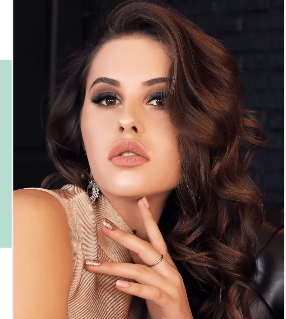 Close-up portrait of a young woman with long, wavy brown hair and striking makeup, featuring bold eyes and soft nude lips. She wears a stylish, sleeveless top and elegant earrings, with one hand resting gently on her face, showcasing manicured nails with a glittery accent. The background is dark, emphasizing her features and the sophisticated atmosphere of the image. Ideal for fashion, beauty, or lifestyle content, this image captures a modern and chic aesthetic.
