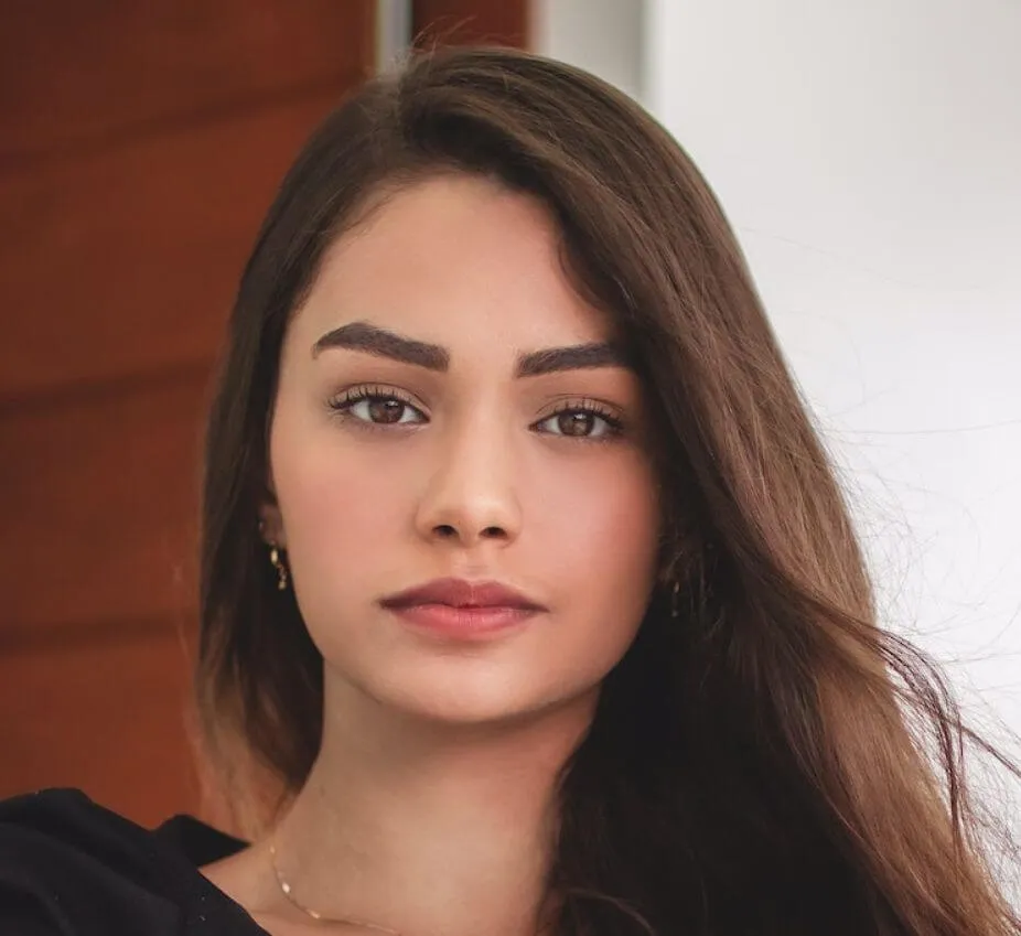 Portrait of a young woman with long, dark hair and natural makeup, looking directly at the camera. She has defined eyebrows and a subtle smile, conveying confidence and approachability. The background features soft, neutral tones that enhance her features, while the lighting is warm and inviting. This image captures a moment of calm and poise, making it suitable for various contexts, including fashion, beauty, or lifestyle content.