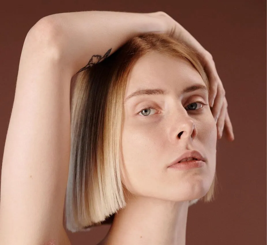 A close-up portrait of a woman with a modern bob hairstyle featuring blonde and dark ombre tones. She gazes confidently at the camera, with her right hand elegantly placed behind her head. The background is a warm brown, enhancing the subject's features. The image captures a blend of contemporary beauty and fashion, suitable for hair styling or beauty-related content. This photograph highlights the model's natural beauty and unique hairstyle, making it ideal for salon promotions or beauty blogs.
