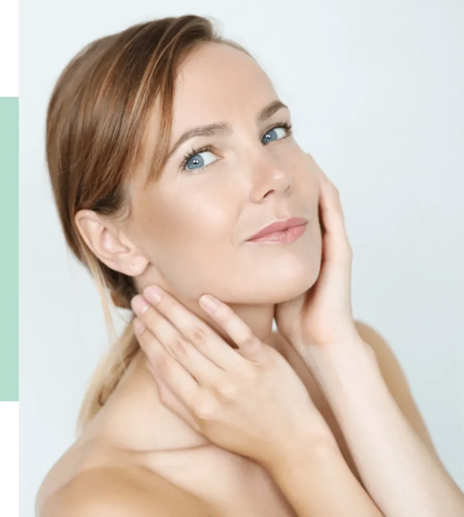 A woman with light brown hair and blue eyes gently holds her face with both hands, showcasing a natural and radiant complexion. The background is softly lit and neutral, enhancing her features. This image conveys themes of beauty, skincare, and self-care, making it ideal for wellness and cosmetic-related content.