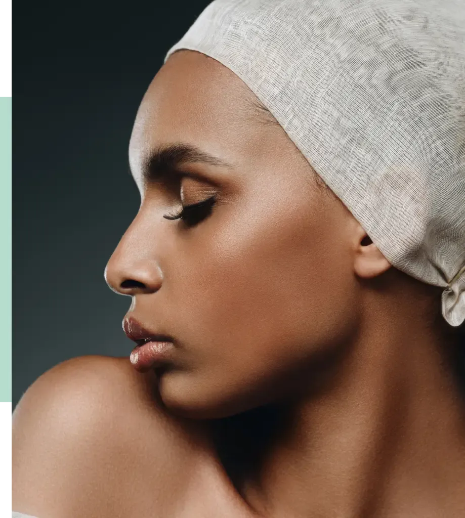 Profile view of a woman with a smooth complexion and defined facial features, wearing a light-colored headscarf. The image highlights her elegant profile against a dark background, showcasing her long eyelashes and full lips. Soft lighting enhances the contours of her face, creating a striking visual effect. This portrait emphasizes beauty and diversity, making it suitable for discussions on fashion, beauty, and cultural representation.