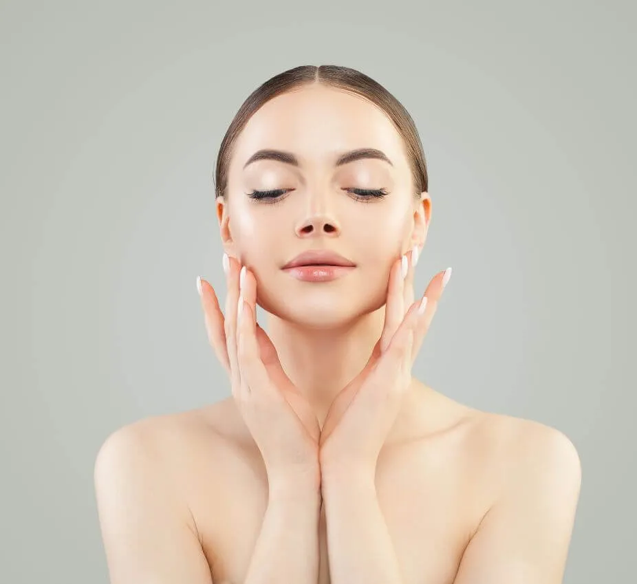 A woman with smooth, radiant skin gently touches her face with both hands, showcasing a natural and serene expression. She has long, straight hair and is featured against a soft gray background, emphasizing her flawless complexion. This image highlights the importance of skincare and self-care routines for achieving healthy skin. Ideal for beauty and skincare blogs, this visual representation promotes confidence and self-love while appealing to audiences interested in beauty tips and products.