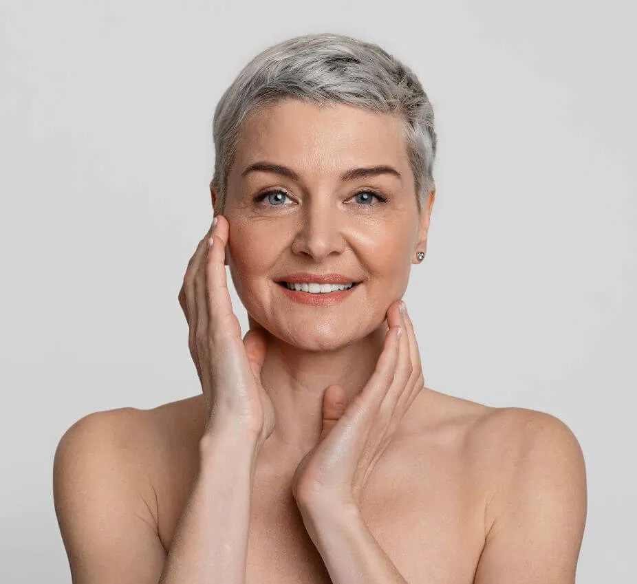 Smiling mature woman with short gray hair gently touching her face, showcasing healthy skin and a natural beauty look. She has blue eyes and a warm smile, conveying confidence and positivity. The background is a soft gray, emphasizing her features. This image represents themes of beauty, aging gracefully, and self-care, appealing to audiences interested in skincare, wellness, and beauty products for mature skin.