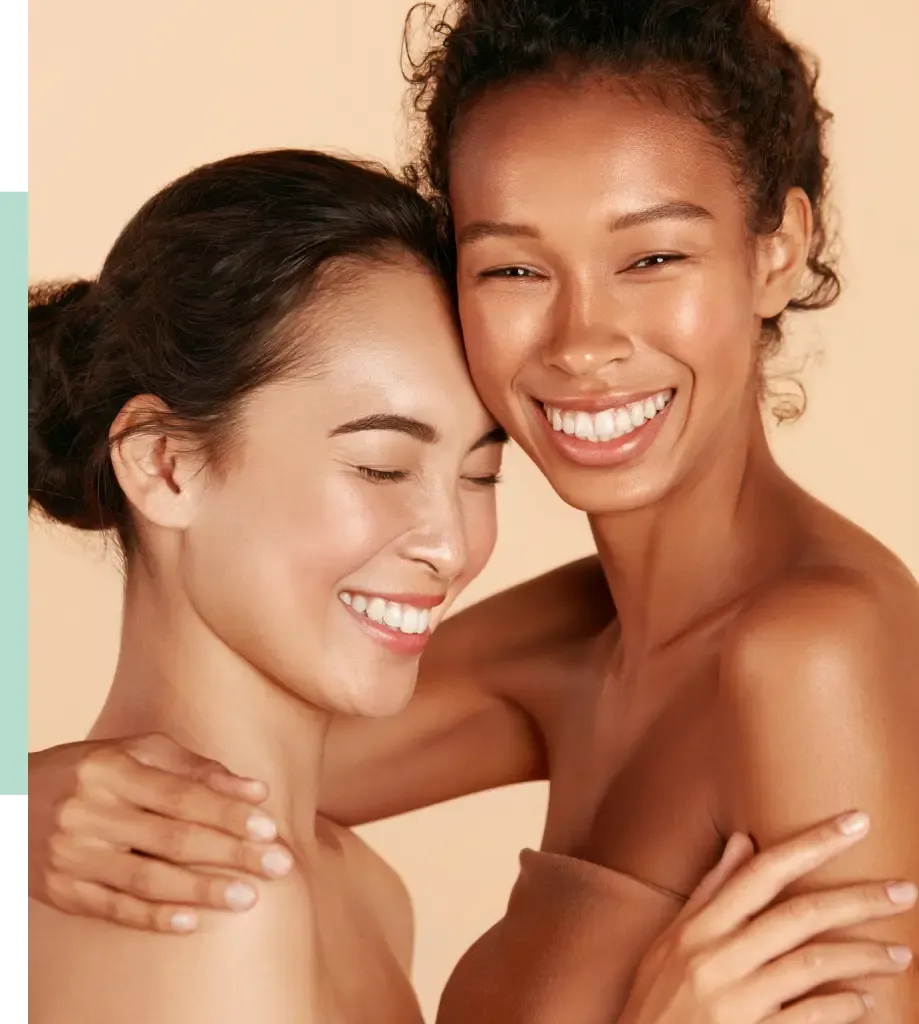Two smiling women embrace each other, showcasing their joyful expressions. One woman has medium-toned skin with dark hair, while the other has a deeper skin tone and curly hair. The background is a soft peach color, enhancing the warm and friendly atmosphere. This image conveys themes of friendship, beauty, and inclusivity, making it suitable for beauty or skincare branding.