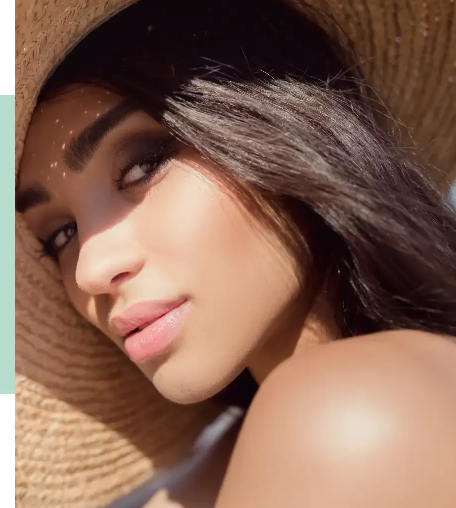 Close-up portrait of a woman with long dark hair, wearing a wide-brimmed straw hat. She has a natural makeup look with soft, glowing skin and a subtle smile. The sunlight casts gentle shadows on her face, highlighting her features. This image captures a serene and confident expression, ideal for beauty and fashion content.