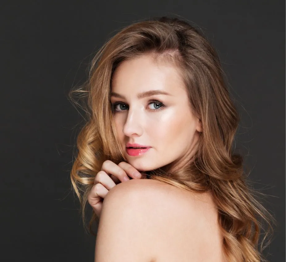 Portrait of a young woman with long, wavy blonde hair and soft makeup, showcasing a natural and elegant look. She is positioned against a dark background, turning her head slightly to the side and looking directly at the camera. Her expression is serene, and she gently holds her hand near her shoulder, emphasizing her graceful pose. The image captures a sense of beauty and confidence, making it suitable for beauty, fashion, or lifestyle themes.