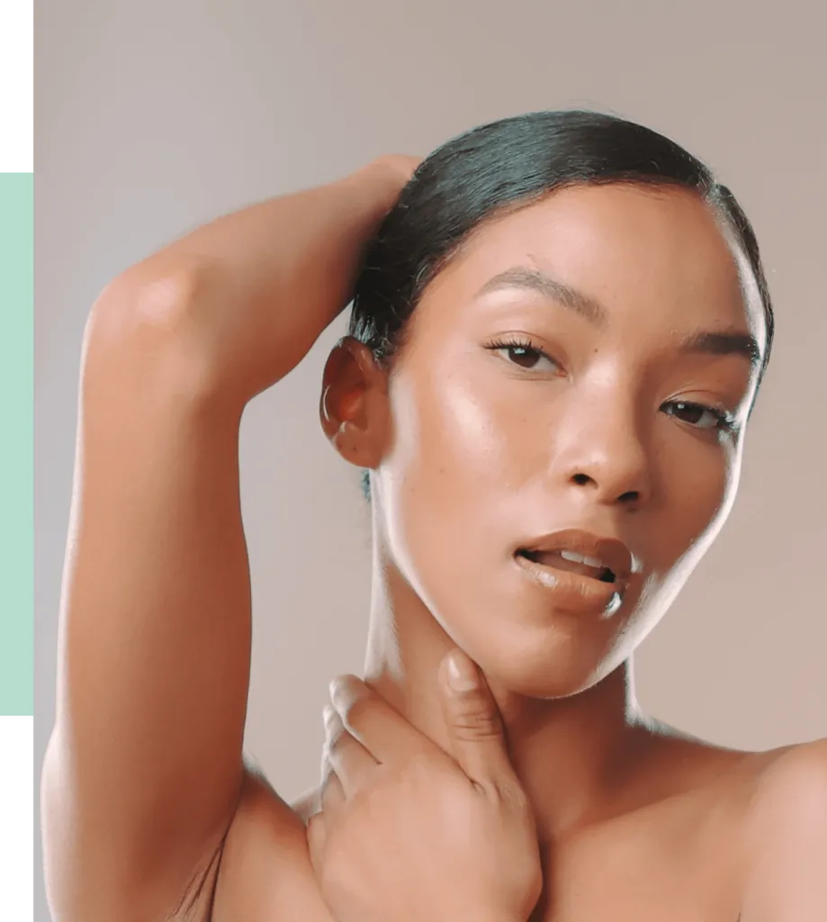 Close-up portrait of a young woman with smooth skin and a sleek hairstyle, showcasing a natural makeup look. She poses with one hand behind her head and the other gently touching her neck, exuding confidence and elegance. The background features soft, neutral tones that enhance her features. This image highlights beauty and self-expression, making it ideal for beauty and skincare content.