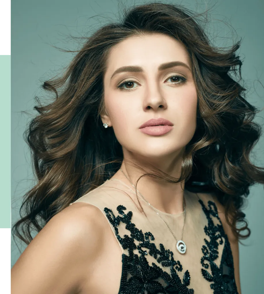 A close-up portrait of a woman with long, wavy brown hair, showcasing her natural beauty. She has a soft makeup look with defined brows and nude lips. The subject wears a black, intricately designed dress with beaded details, complemented by a delicate silver necklace featuring a circular pendant. The background is softly blurred, enhancing her striking features and elegant pose. This image captures a sophisticated and confident appearance, ideal for beauty and fashion content.