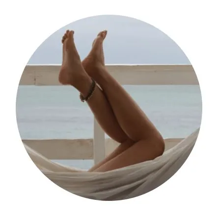 A close-up image of a person's legs relaxing in a hammock, with the ocean in the background. The legs are crossed at the ankles, showcasing smooth skin and a bracelet on the ankle. The scene conveys a sense of tranquility and leisure, perfect for a beach vacation or relaxation theme. The soft, muted colors of the ocean and sky create a calming atmosphere, enhancing the overall peaceful vibe of the moment. This image represents the idea of unwinding and enjoying nature, ideal for travel or wellness-related content