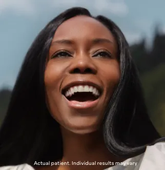 Smiling woman with long black hair outdoors, showcasing a joyful expression against a natural backdrop. This image conveys a sense of happiness and confidence, suggesting positive outcomes related to personal care or health treatments. The text at the bottom notes that the individual is an actual patient and results may vary, emphasizing authenticity. Ideal for health and wellness content, this image promotes positivity and self-improvement.