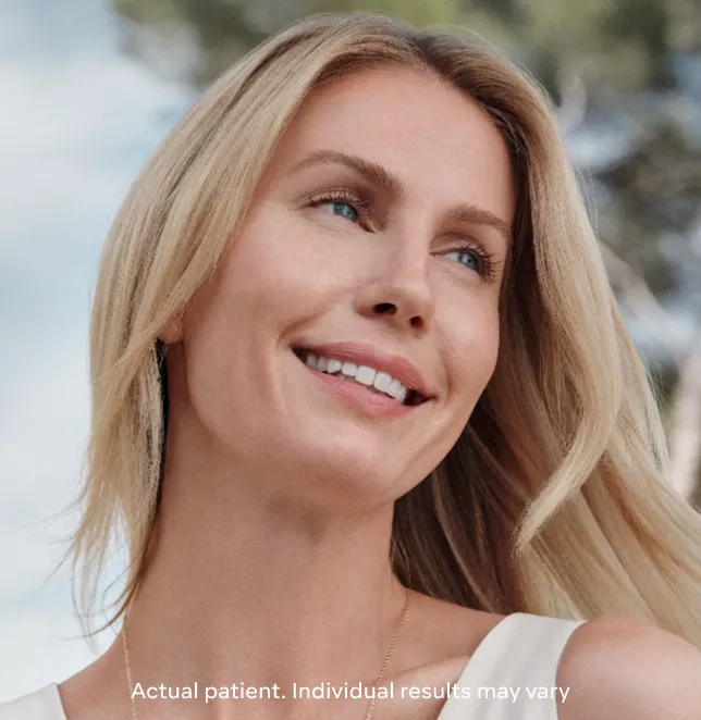 Smiling woman with long blonde hair outdoors, showcasing healthy skin and a natural glow. The background features soft greenery, enhancing the serene atmosphere. This image represents the positive outcomes of aesthetic treatments, emphasizing individual results may vary. Ideal for promoting beauty and wellness services.